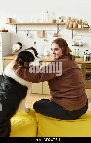 A young woman sits on a bright couch, happily interacting with her dog in a warm kitchen. Stock Photo