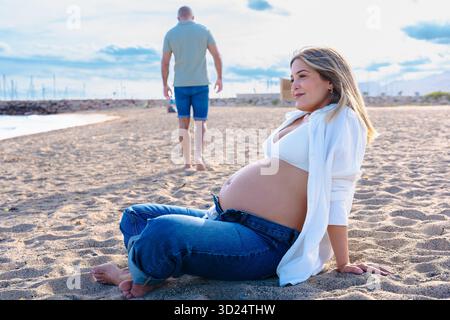 Pregnant woman on beach, sitting on sand, watching man walking away near sea. Stock Photo