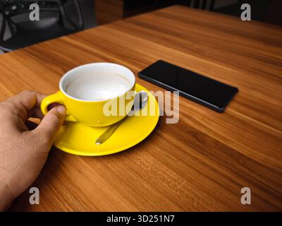 Close-up of hand holding a yellow coffee cup with spoon and smartphone on wooden table, minimalistic cafe concept with warm tones and modern lifestyle Stock Photo