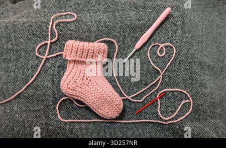 Handcrafted pink baby sock with crochet hook and yarn heart on gray knitted fabric background. Stock Photo