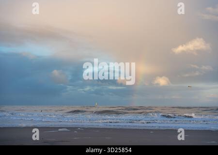 A faint rainbow glows through pastel clouds over the rolling sea. Gentle waves meet the quiet shore in soft evening light. Stock Photo