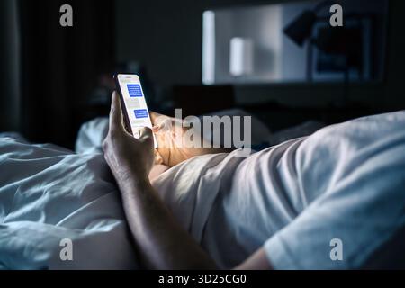 Phone in bed. Text message. Man texting. Screen time at night. Using smartphone messenger app in dark bedroom or hotel room before sleeping. Stock Photo