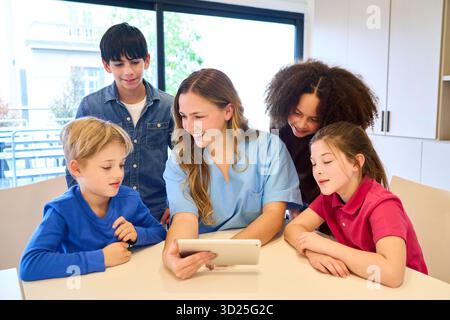 A friendly clinician in blue scrubs leads a diverse group of children, using a tablet to guide engaging rehabilitation activities in a pediatric setti Stock Photo