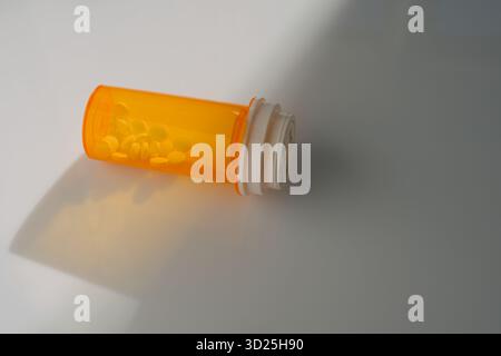 Single orange prescription medicine bottle lying on its side on a white surface with a few yellow pills visible inside. Stock Photo