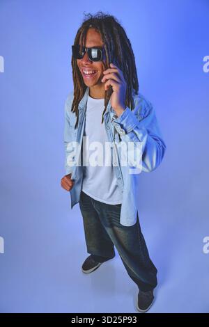 Smiling young man with dreadlocks wearing sunglasses, talking happily on a mobile phone call Stock Photo