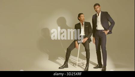 Posing male colleagues wearing tailored suits in studio, with white stool and wristwatch Stock Photo