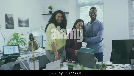 Smiling three coworkers wearing work attire standing at workstation in office, with laptop monitors Stock Photo