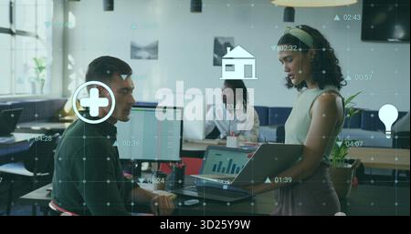 Holding laptop, office worker discussing data with coworker in open-plan office, digital icons Stock Photo