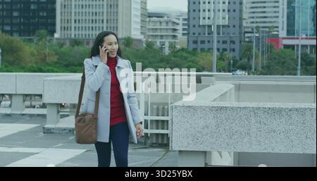 Walking woman wearing gray coat talking on smartphone at city plaza, with railing and brown bag Stock Photo