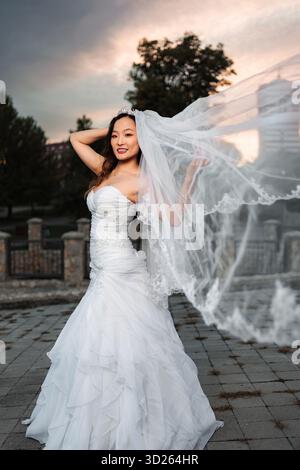 Chinese bride smiling in flowing wedding dress and veil at sunset Stock Photo