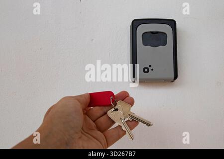 Person holding silver house keys with red keychain near a secure wall mounted lock box. Stock Photo