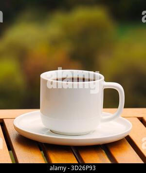 A white coffee cup with a brown liquid in it sits on a wooden table. The cup is placed on a white saucer, which is also on the table. Concept of relax Stock Photo