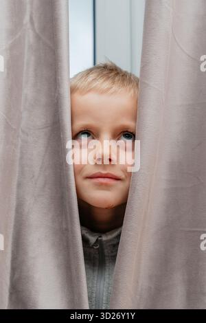 Child looks up through soft curtains, face framed by gentle light dreamy, contemplative indoor portrait Stock Photo