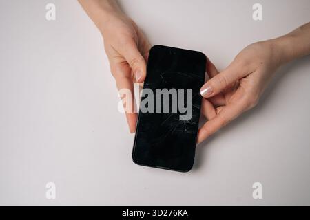 Top view of woman removing broken tempered glass screen protector from smartphone, revealing cracked protection film on white table. Stock Photo