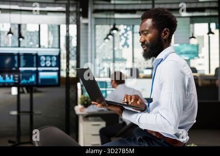 African American male manager sits on table in corporate office, reviewing emails, online data and business documents. Black man holds laptop, planning and managing financial projects in workplace. Stock Photo