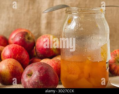 A glass jar of homemade apple jam or honey, with a spoon, surrounded by fresh red apples Stock Photo