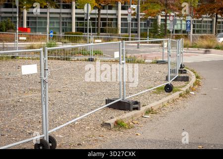 A fence secures a vacant lot in a busy urban location. The scene shows a gray path along with autumn leaves scattered on the ground and modern buildin Stock Photo