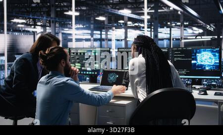 Teamworking data center technicians using machine learning algorithms to analyze large datasets. Server room colleagues gathered together at desk, collaborating, using AI neural networks, camera A Stock Photo