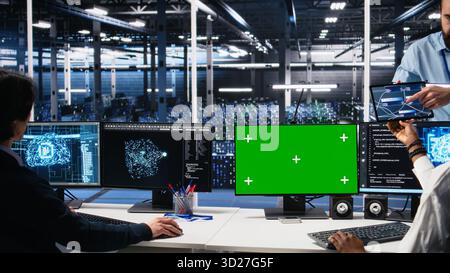 Data center manager overseeing team of technicians managing AI systems using green screen PC. Supervisor checking work of teamworking server hub colleagues using mockup computer, camera A Stock Photo