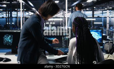 Multiracial technicians in diverse data center review AI diagnostics, analyzing neural network performance. Asian man and african american woman optimize artificial intelligence systems, camera A Stock Photo