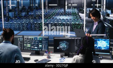 Software developers cooperating in data center, reviewing AI diagnostics on computer. IT staff members discussing in server room, optimizing machine learning systems, camera A Stock Photo