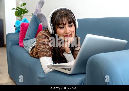 Woman enjoying video content or music at home on sofa with headphones and laptop Stock Photo