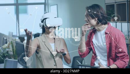 Gesturing Chinese woman using VR headset while colleague using tablet at office with wireless icons Stock Photo