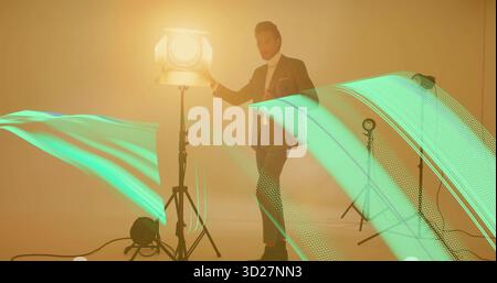 Posing Asian man wearing dark suit leaning near studio spotlight in studio, with green LED trails Stock Photo