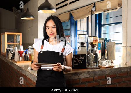 Asian Woman Barista Wearing Black Apron Looking at Camera and Holding Tablet, Standing Next to Cafe Bar Stock Photo