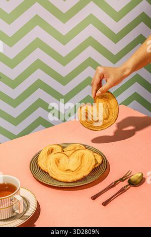 Heart-Shaped Palmier Cookies with Hand Lifting One on Pink Table and Tea Cup in Chevron Background Stock Photo