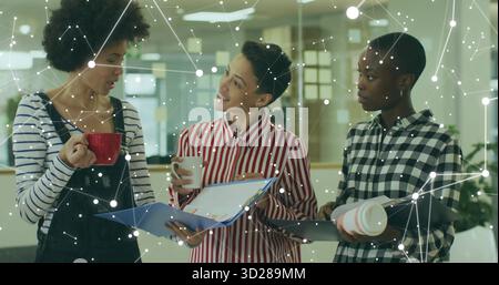 Discussing three women holding coffee mugs reviewing folders in modern office, with digital overlay Stock Photo