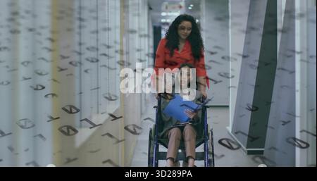 Discussing seated woman gesturing at blue folder on wheelchair in corporate hallway, with colleague Stock Photo