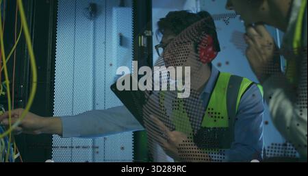 Male engineer with vest holding clipboard in data center rack, colleague adjusting patch cables Stock Photo