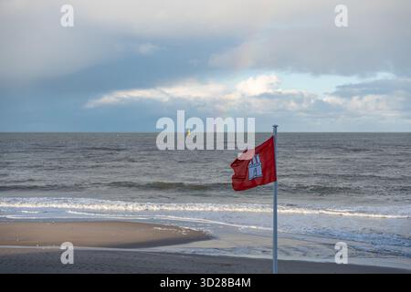 Red flag with castle emblem fluttering by the sea under a cloudy sky. A faint rainbow appears over the ocean horizon. Stock Photo