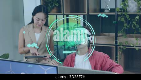 Pointing woman holding tablet and guiding man wearing red jacket, VR headset at office desk Stock Photo