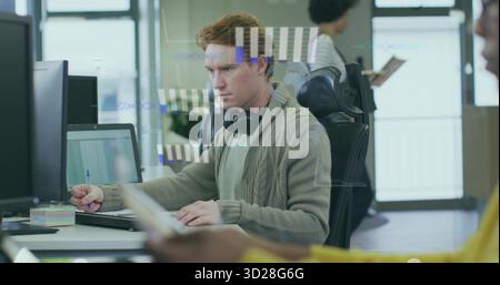 Typing man with headphones working at office desk, with laptop, monitor and sticky notes Stock Photo