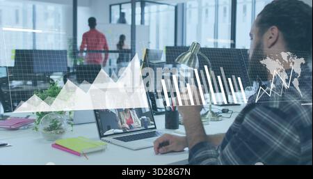 Gesturing male office worker using laptop in open plan office with graphs overlay, copy space Stock Photo