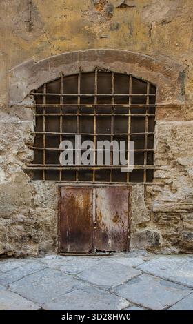 A barred window in Albergheria quarter of Palermo, Italy. The wall features rough stone blocks at the base & stucco on the upper sections. Stock Photo