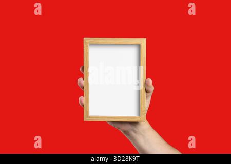 A Caucasian man's hand holds a blank wooden picture frame with empty white copy space for a mockup, isolated on a solid red background. Stock Photo