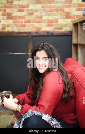 Smiling woman with tinsel holding festive mug on red sofa indoor Stock Photo