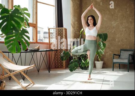 Fit smiling woman practicing tree yoga pose in cozy place Stock Photo