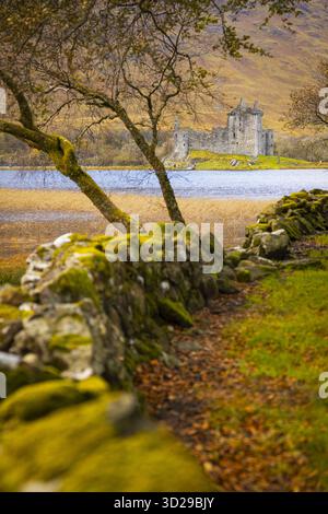 View of ancient stone wall covered with vibrant green moss leads the eye toward a tranquil loch and imposing castle ruins, Isle of Skye, Scotland, Uni Stock Photo