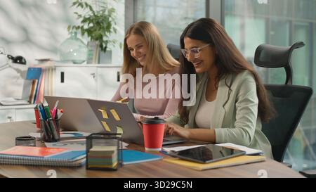 Two multiethnic women discuss graphic statistics corporate growth in office diverse businesswomen informal business girls Caucasian Indian ladies Stock Photo