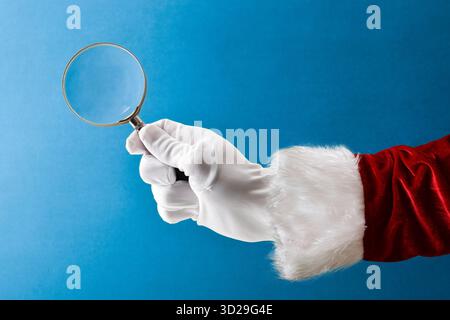 Santa Claus in suit and glove holding magnifying glass isolated on blue background Stock Photo
