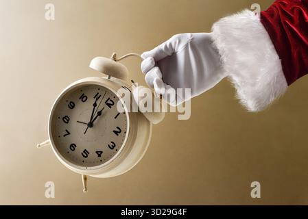 Santa Claus hand in suit and gloves holding an alarm clock on New Year's Eve showing countdown on brown isolated background. Stock Photo