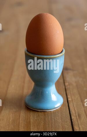 Brown egg resting in a vibrant blue ceramic egg cup, presented on a light wooden table. Healthy breakfast food concept Stock Photo