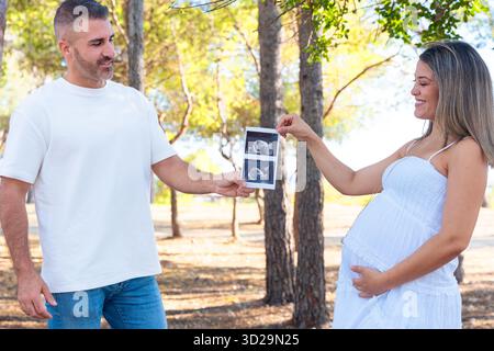 Pregnant couple holding ultrasound photo together outdoors in sunny park Stock Photo