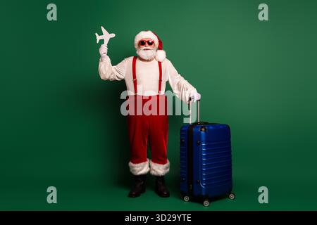Santa in red costume with hat and beard posing beside a blue suitcase holding a plane cutout for Christmas travel and holiday celebration on a green Stock Photo