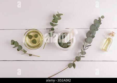 Natural eucalyptus oil in a bowl and glass bottle, mortar and pestle and fresh eucalyptus leaves on a white wooden table. top view Stock Photo