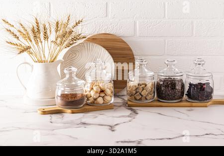 A set of glass jars with various bulk products stand on wooden boards and a marble white countertop. compact storage of products Stock Photo